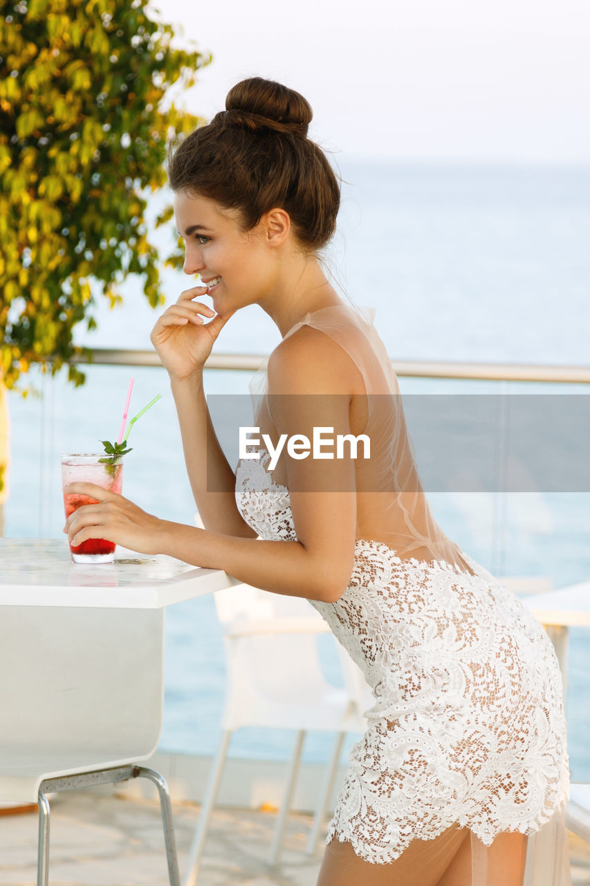 Beautiful woman holding cocktail while sitting at restaurant against sea