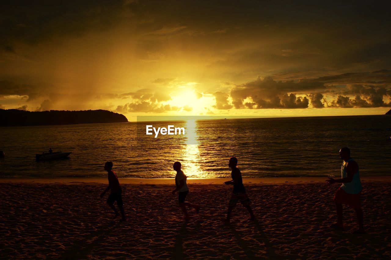 People walking on sand at beach against sunset sky