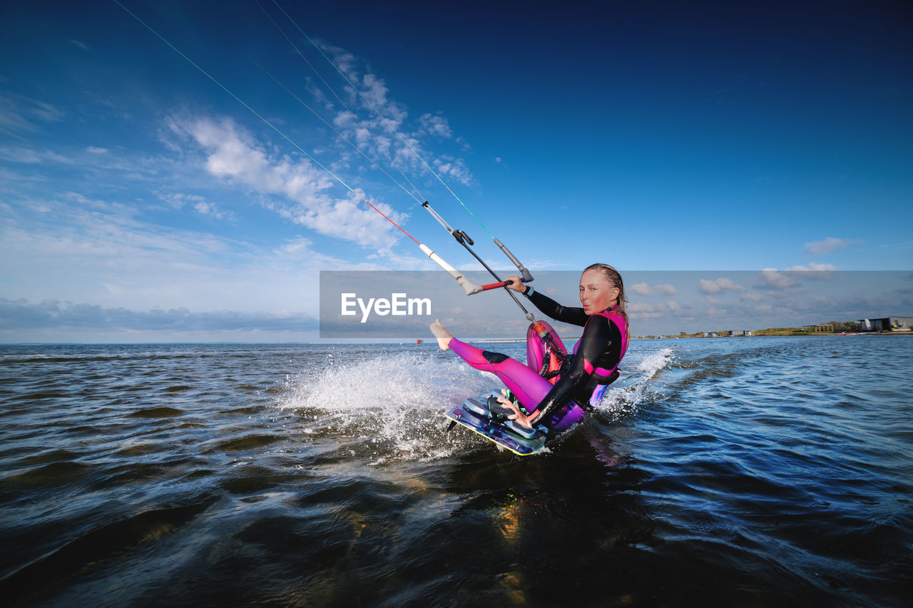 A young caucasian woman in a wetsuit rides a kiteboard. an athlete performs a trick while sitting on