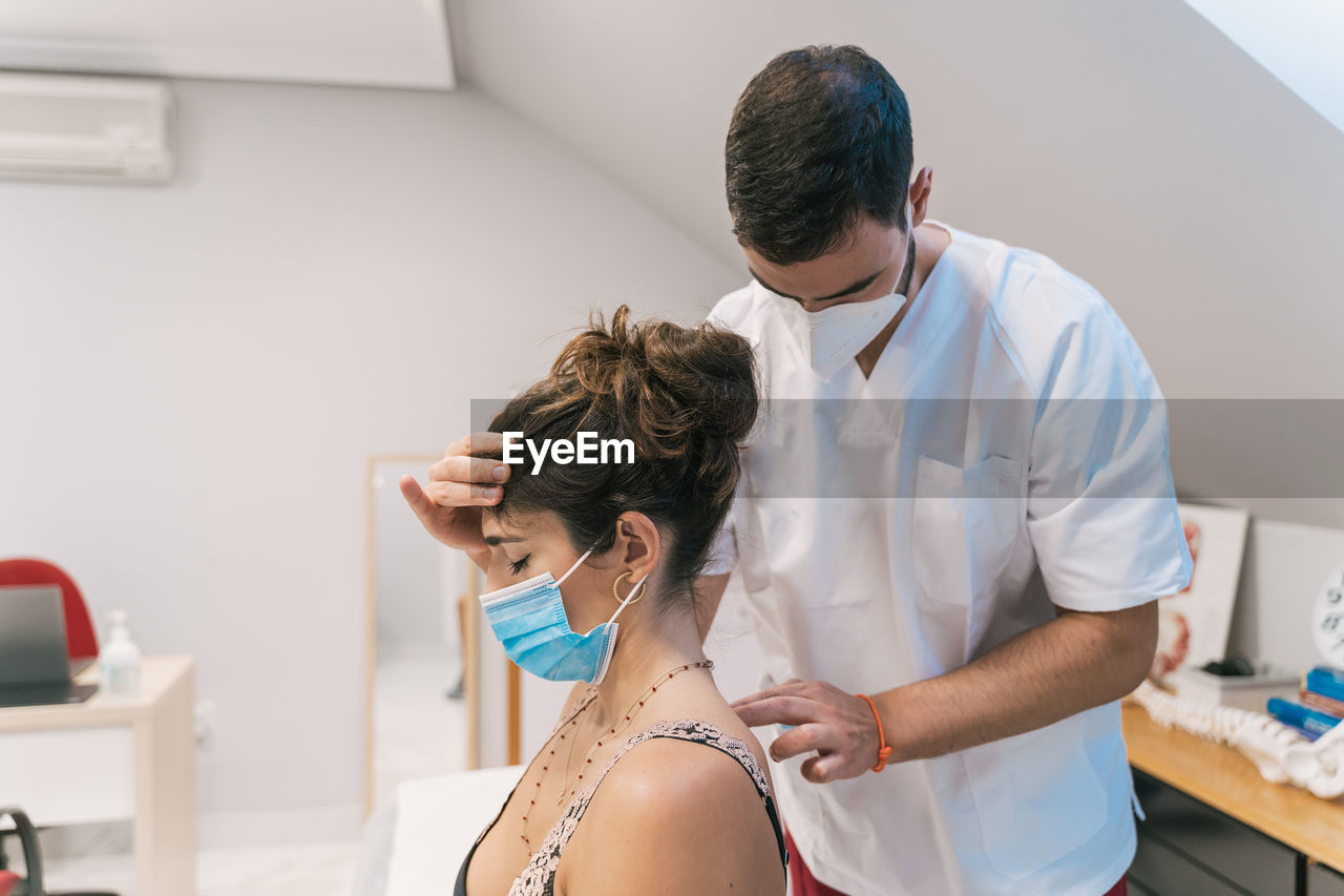 High angle of male physiotherapist massaging back of calm female patient in mask sitting on medical table in modern clinic