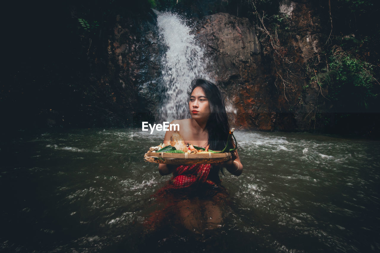 Young woman holding food in wicker basket against waterfall