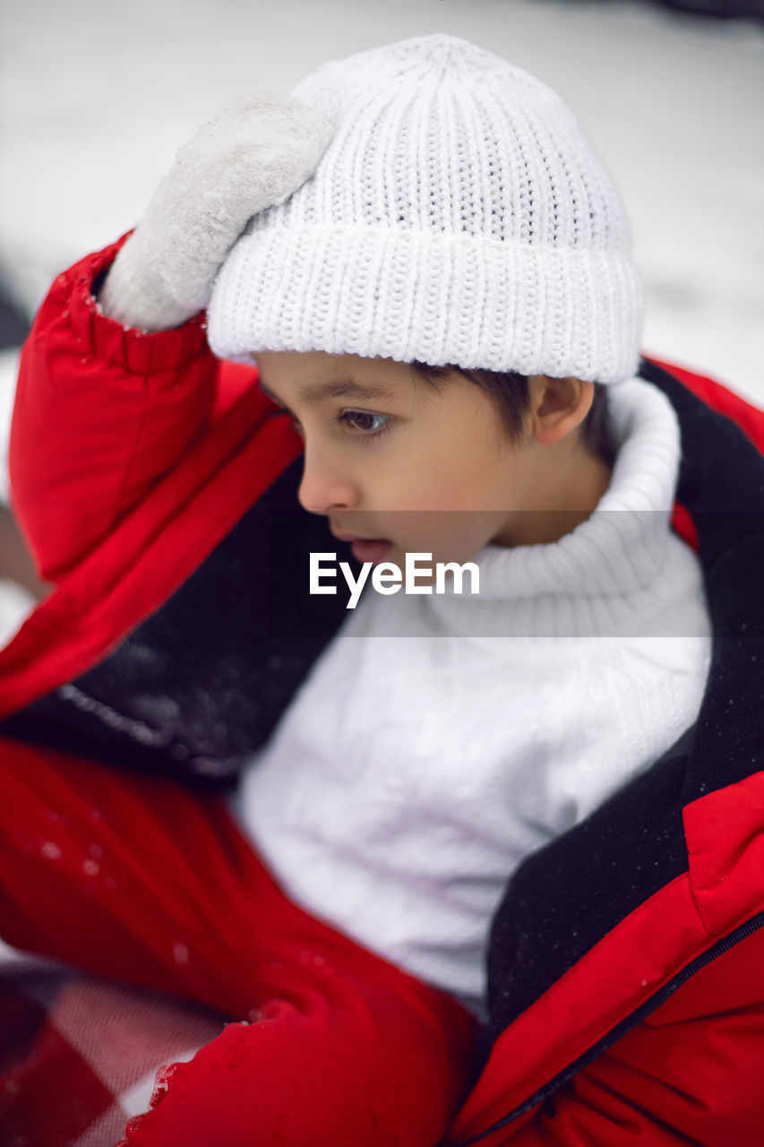 A boy child in a red jacket, hat and mittens sits outside the house on christmas day in winter