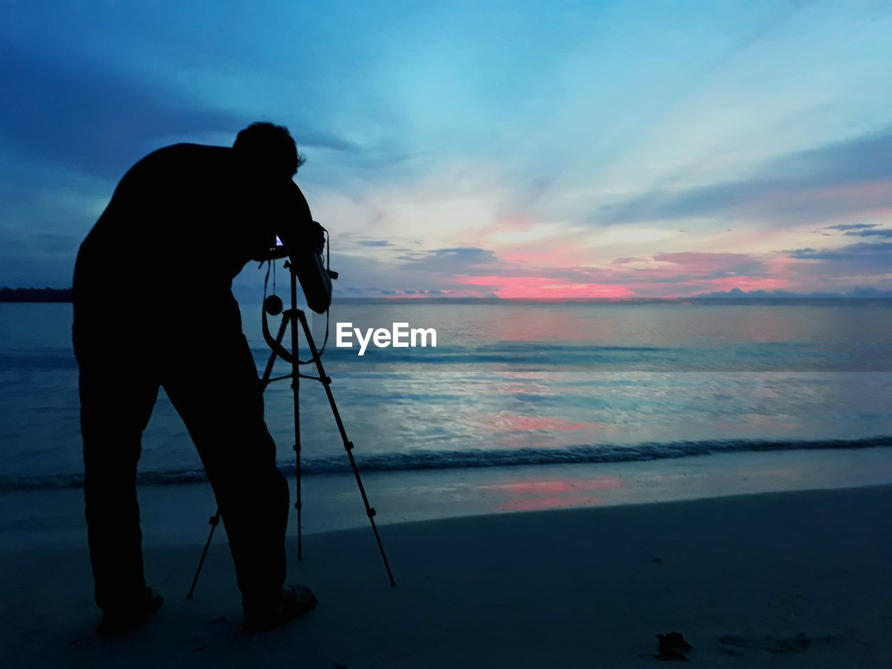 Rear view of man photographing while standing on shore at beach during sunset