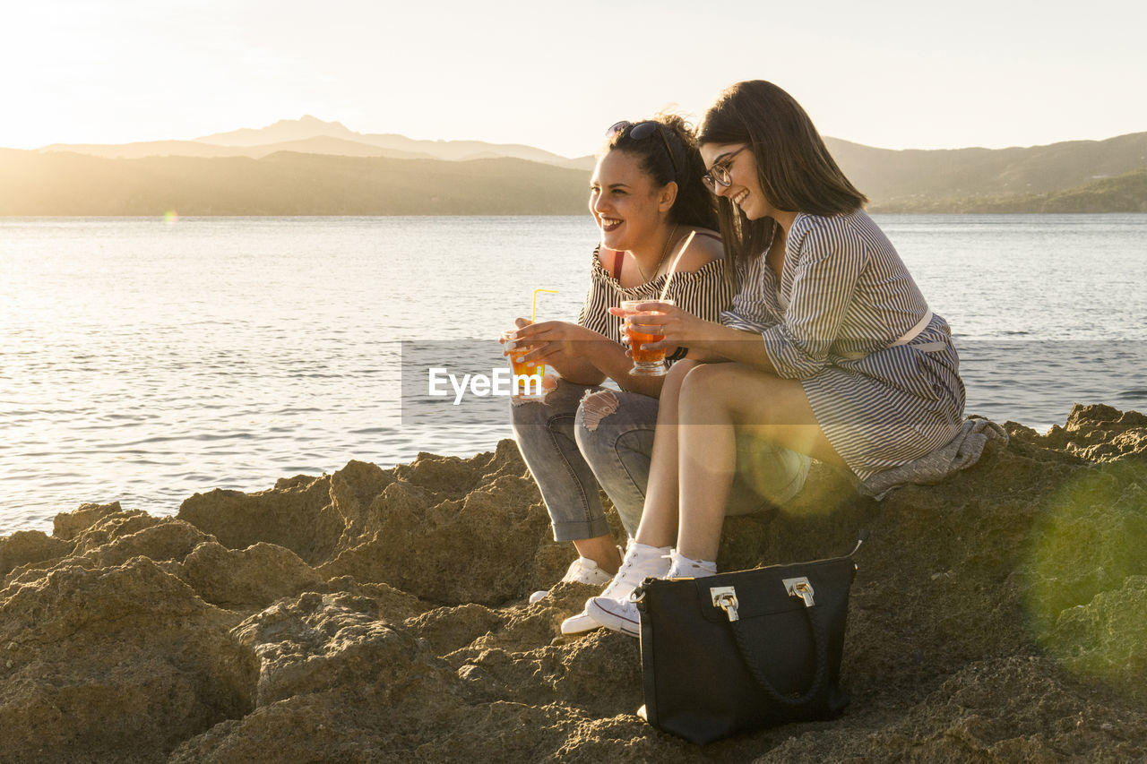 Full length of female friends having drink while sitting on rocks at beach