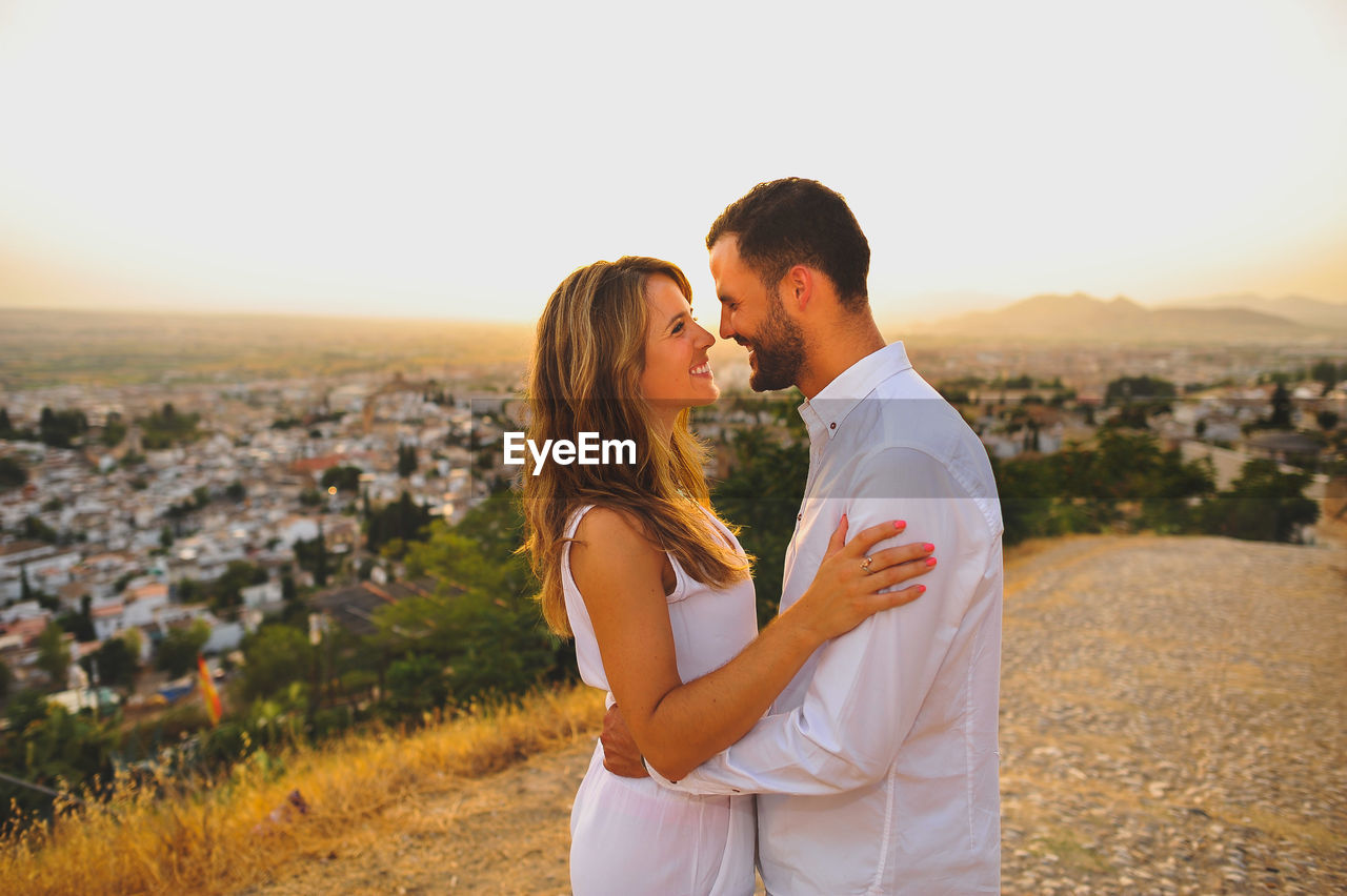 Happy romantic couple standing on cobbled road against sky during sunset