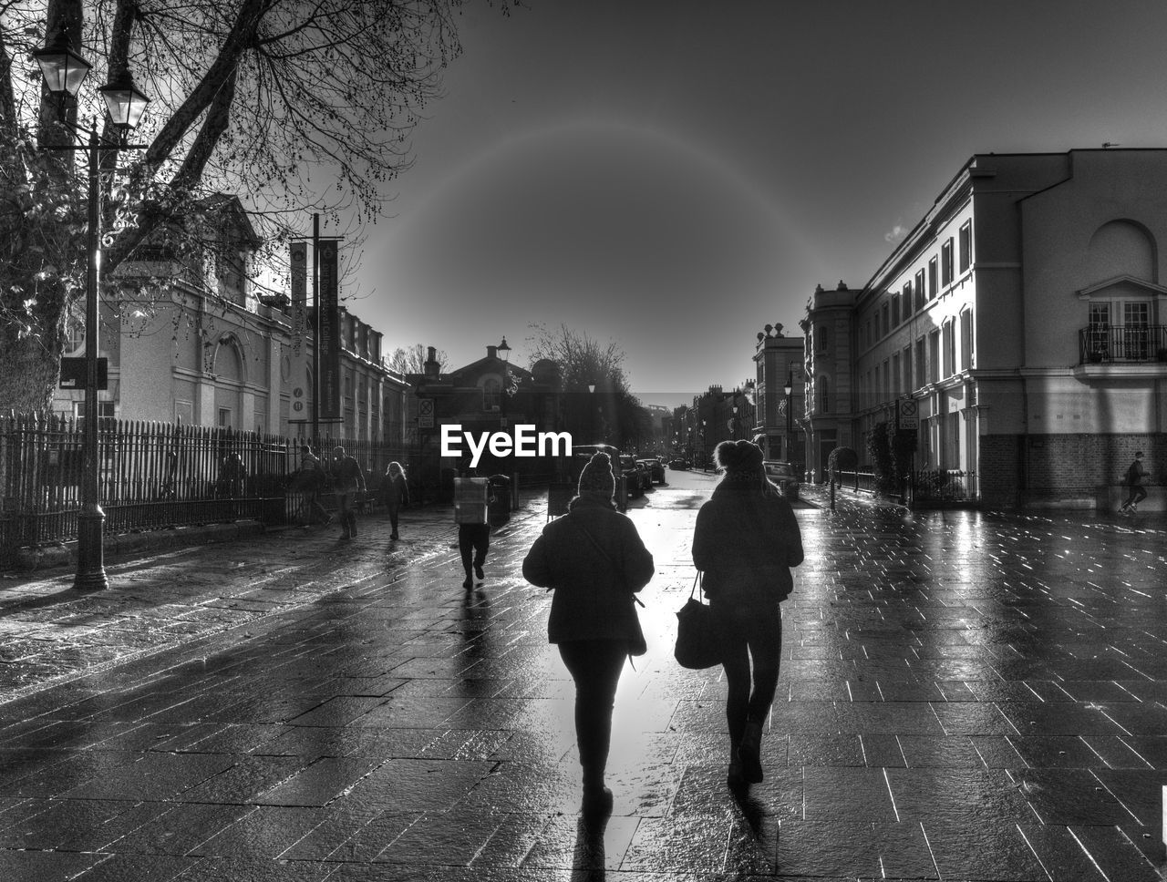 REAR VIEW OF WOMEN WALKING ON WET STREET AMIDST BUILDINGS