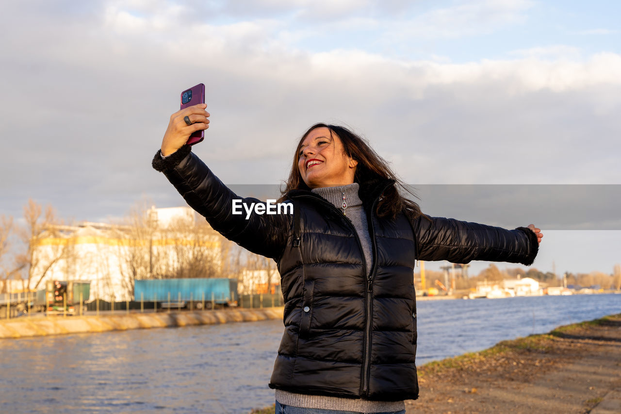 Middle aged woman wearing winter clothes taking selfie by a river - concept of people in recreation