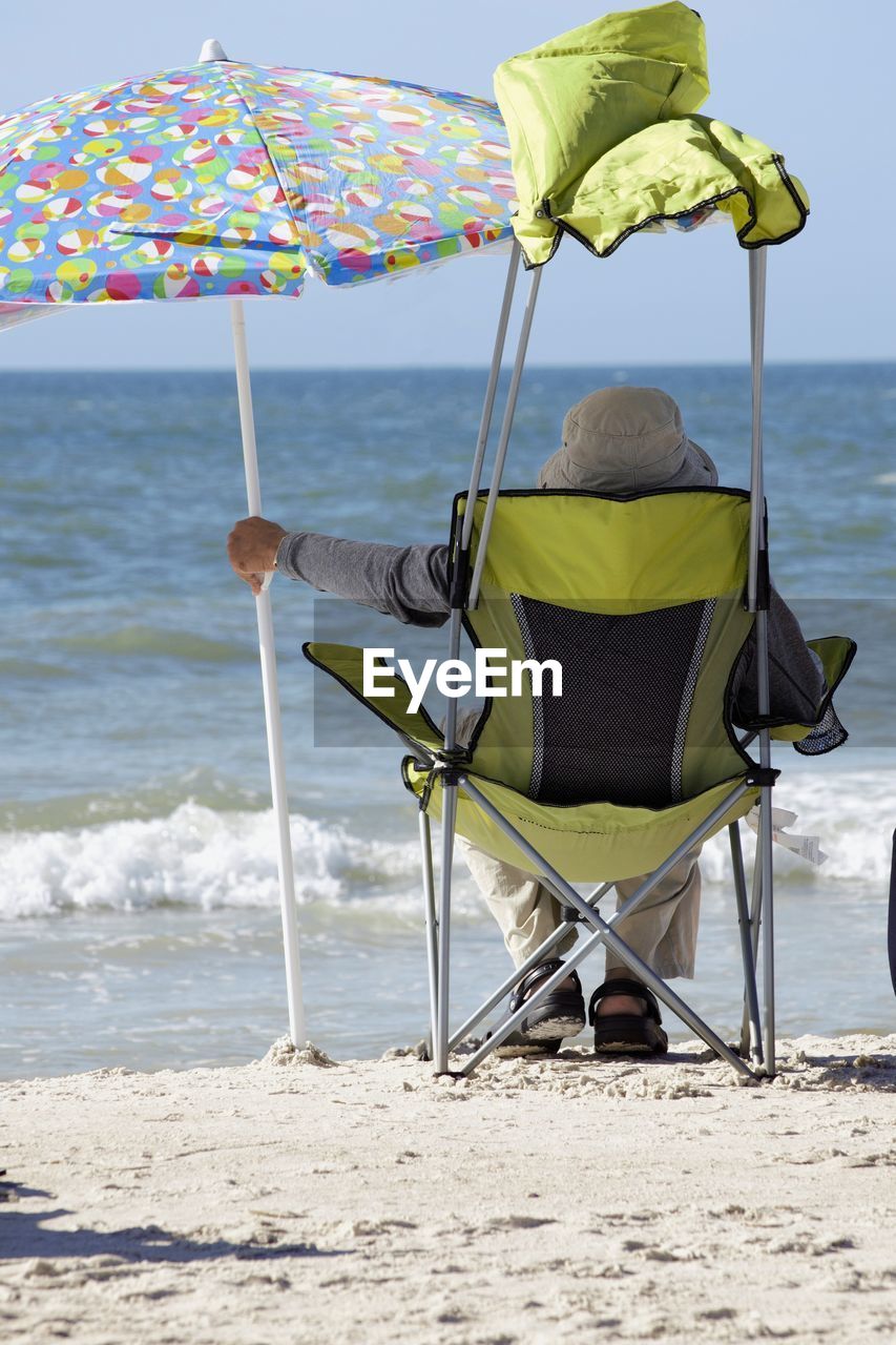 A rear view of a senior sitting on a florida beach holding an umbrella looking at waves.
