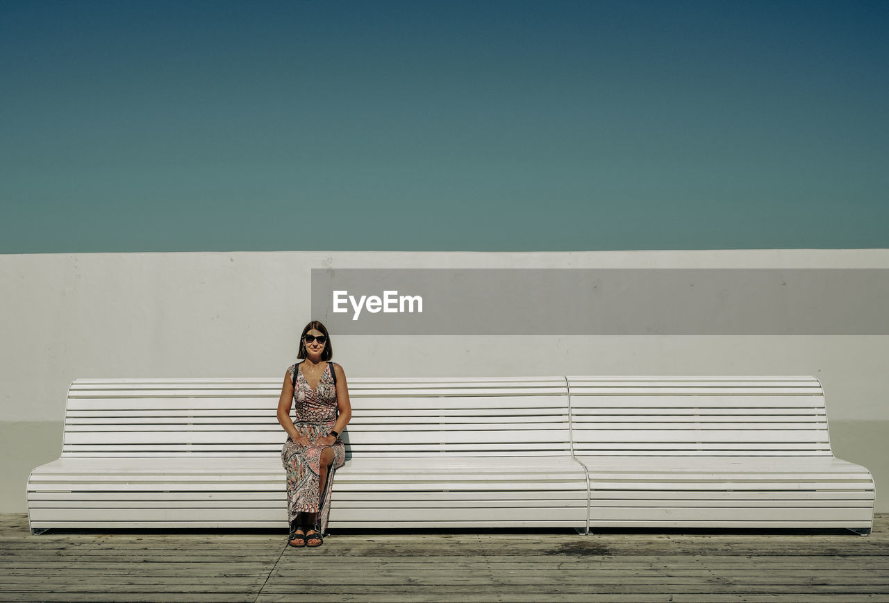 A lonely woman sits on a white bench with a white background and the sky behind her