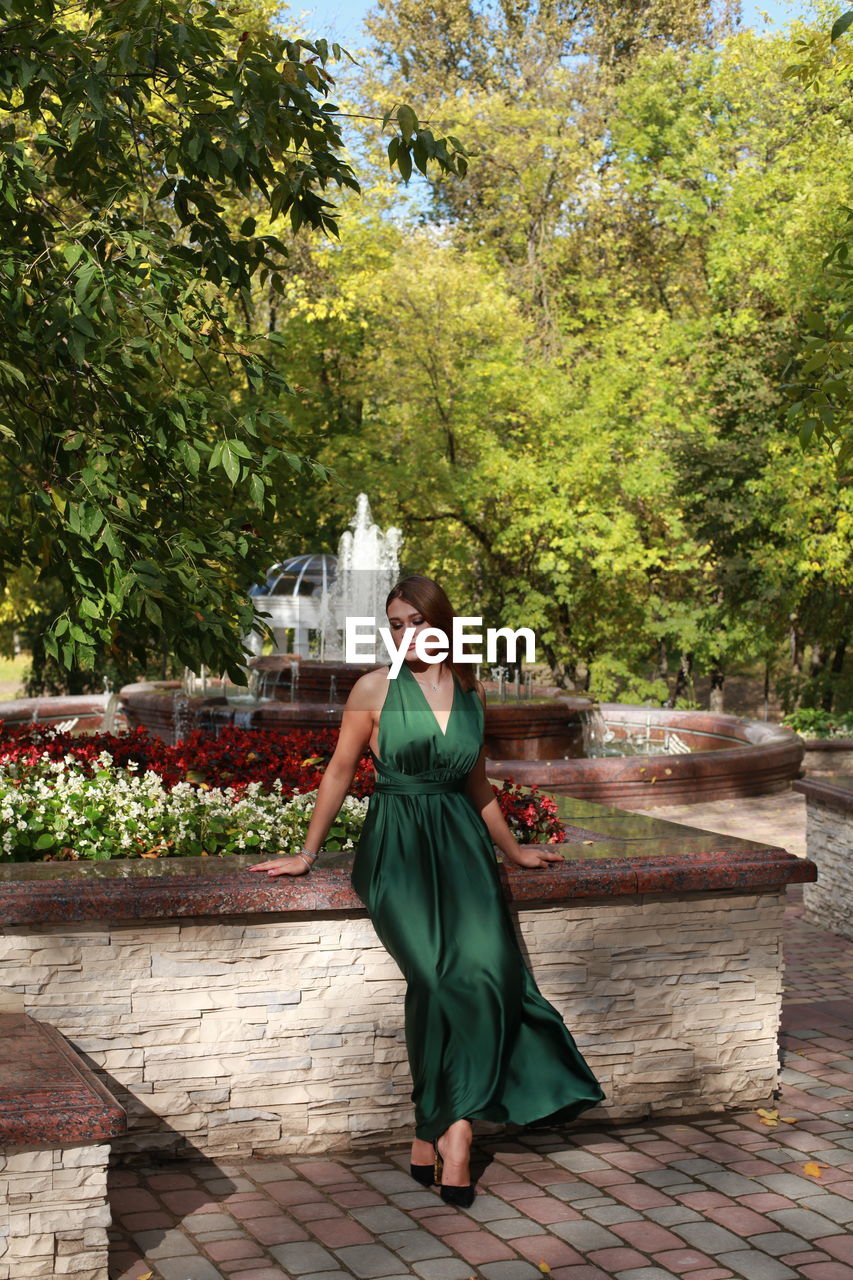 Portrait of woman sitting by tree against autumn park background, wearing green silk dress 