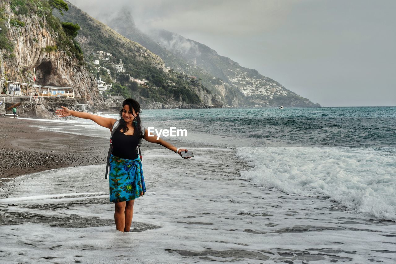 Young woman with arms outstretched standing at beach against cloudy sky