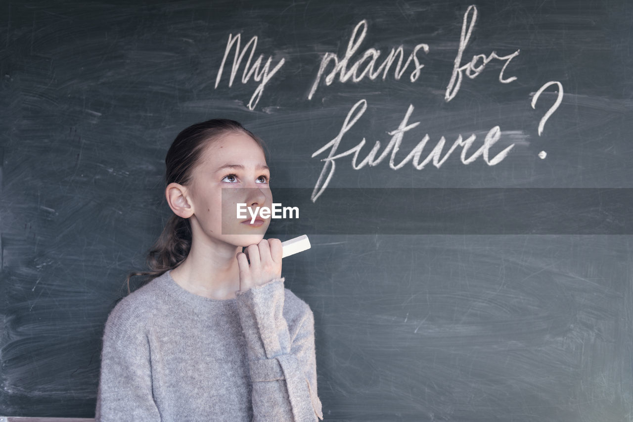 Girl looking away while standing by blackboard with text in classroom