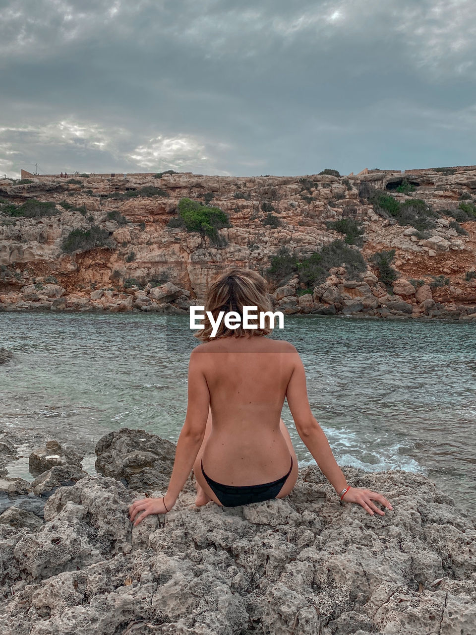 Rear view of shirtless woman sitting on rock against sky