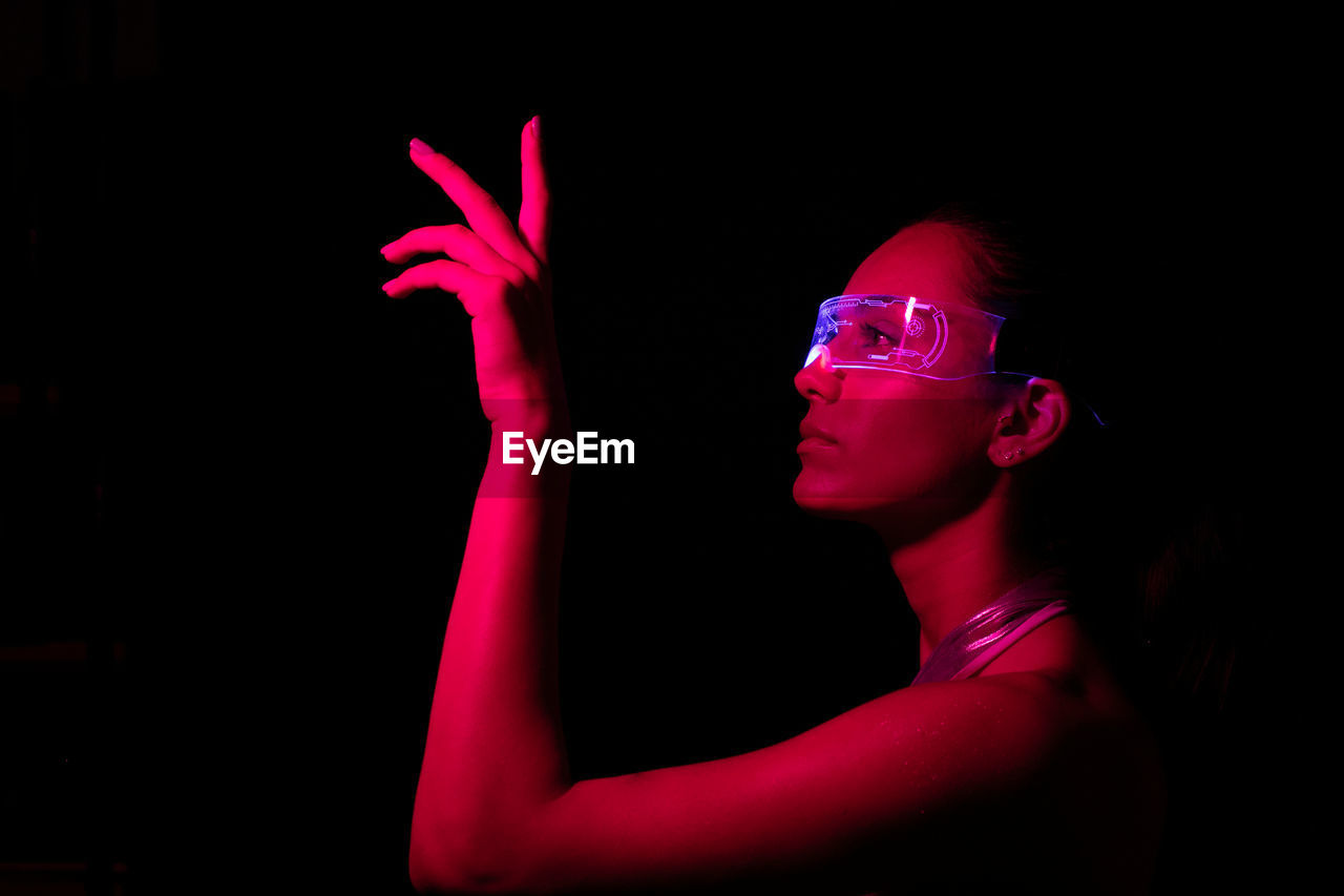 Side view of serious focused young female touching invisible object while experiencing virtual reality in modern transparent vr glasses in dark studio with bright pink neon illumination