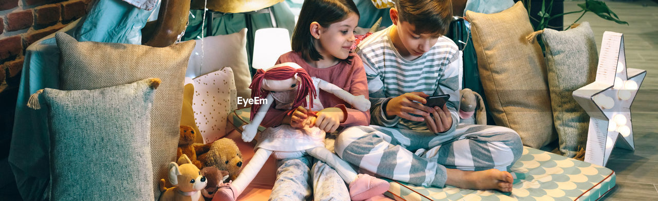 Banner of happy little girl embracing rag doll while looking boy playing game with phone sitting