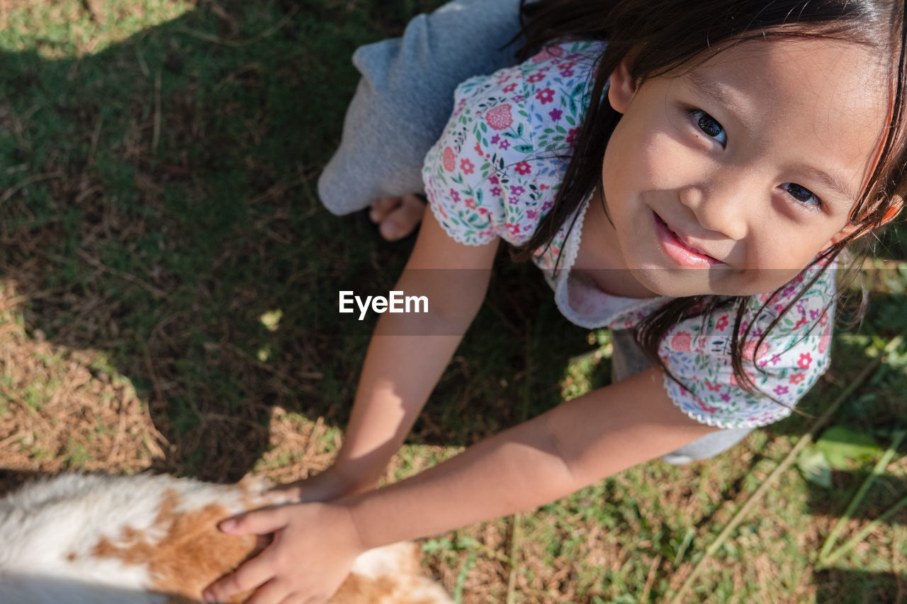 High angle portrait of cute smiling girl holding cat while crouching on grassy field