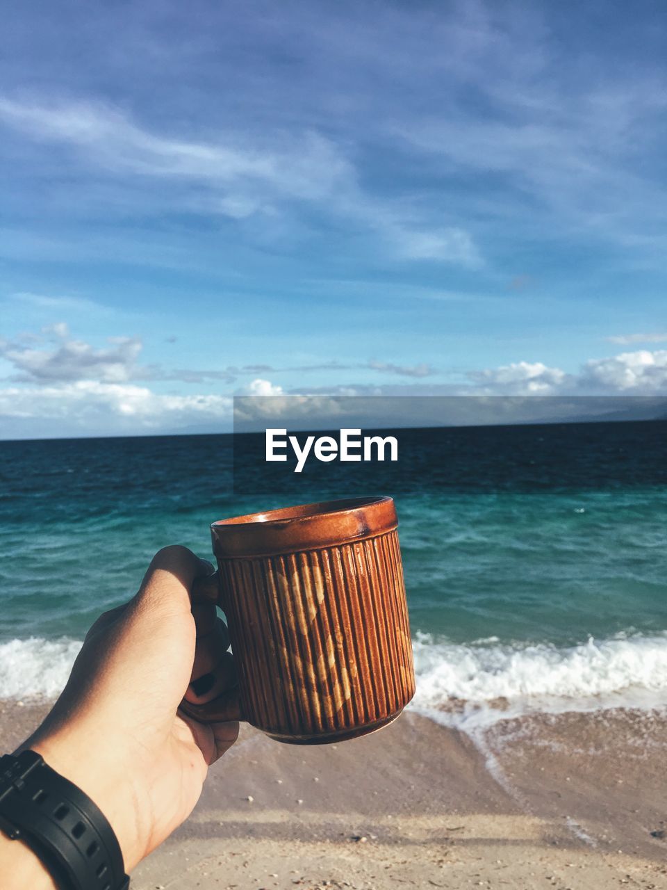 Cropped hand holding coffee cup at beach against sky