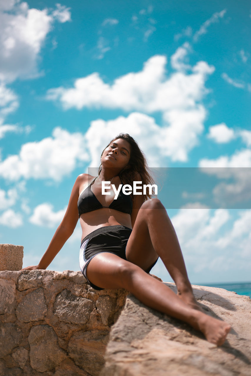 Portrait of woman in bikini sitting on rock against sky