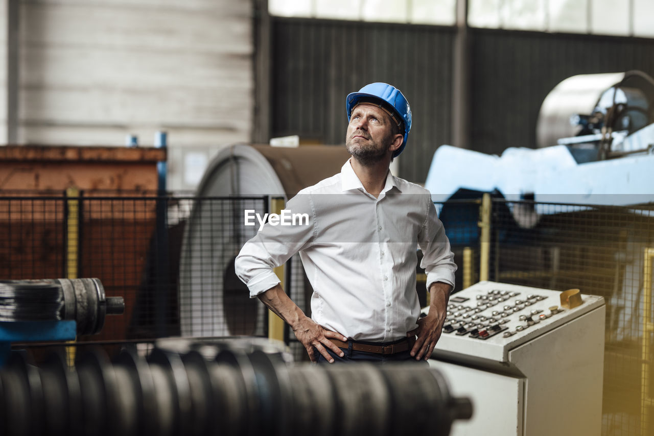 Male entrepreneur with hand on hip looking up while standing by control panel in factory