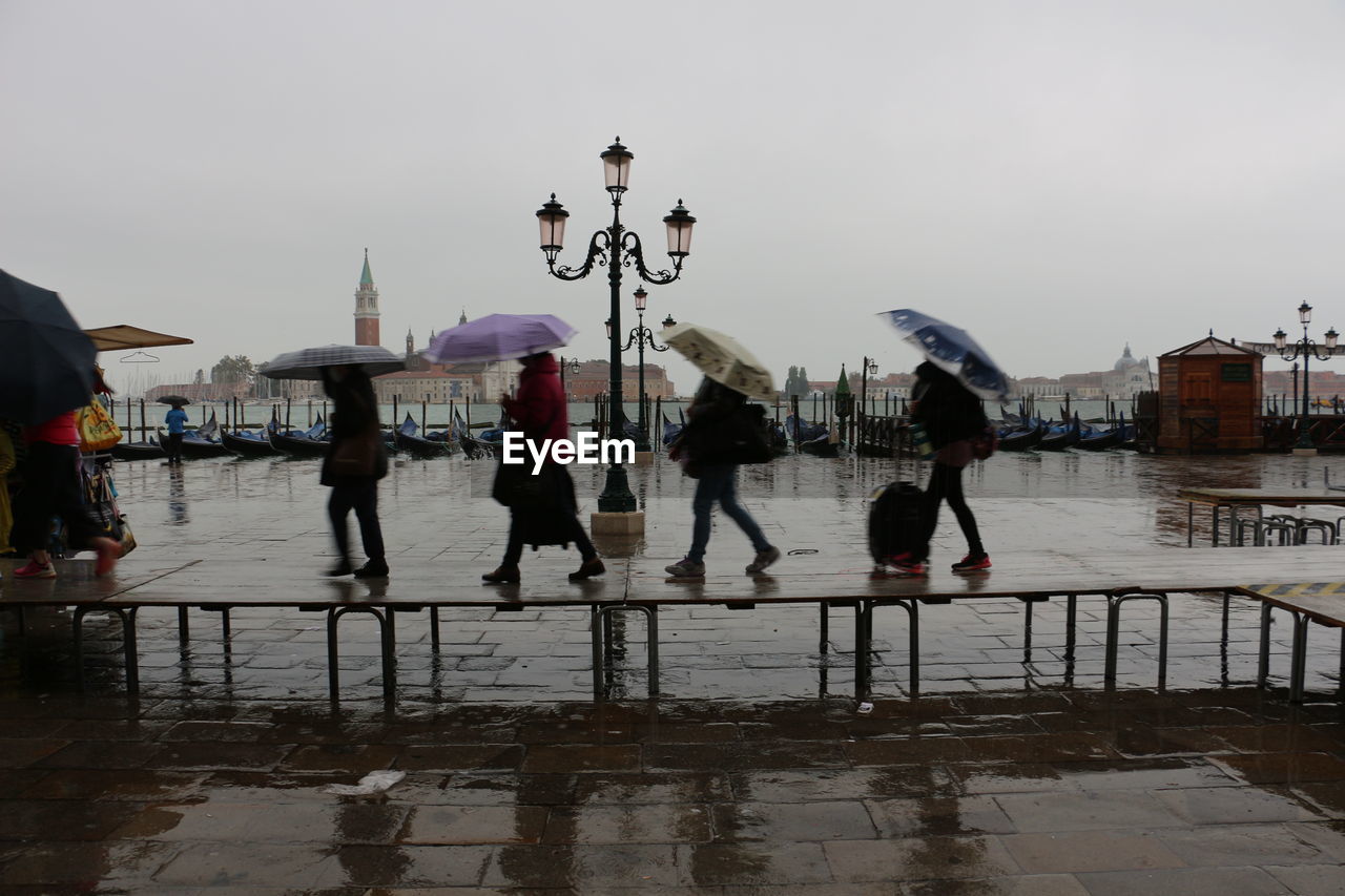 People with umbrellas walking on wet promenade against sky in city