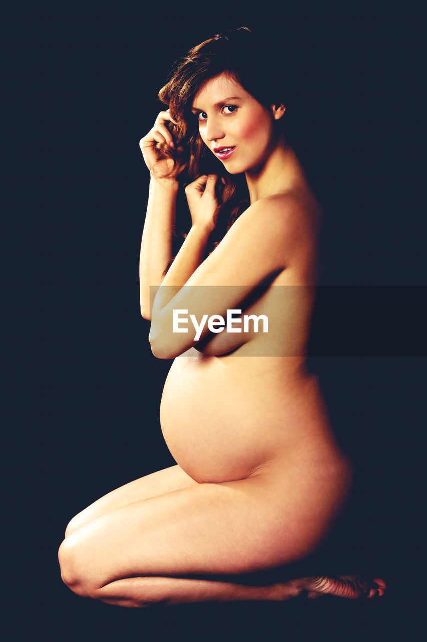 Portrait of naked pregnant young woman covering breasts with hand in hair against black background