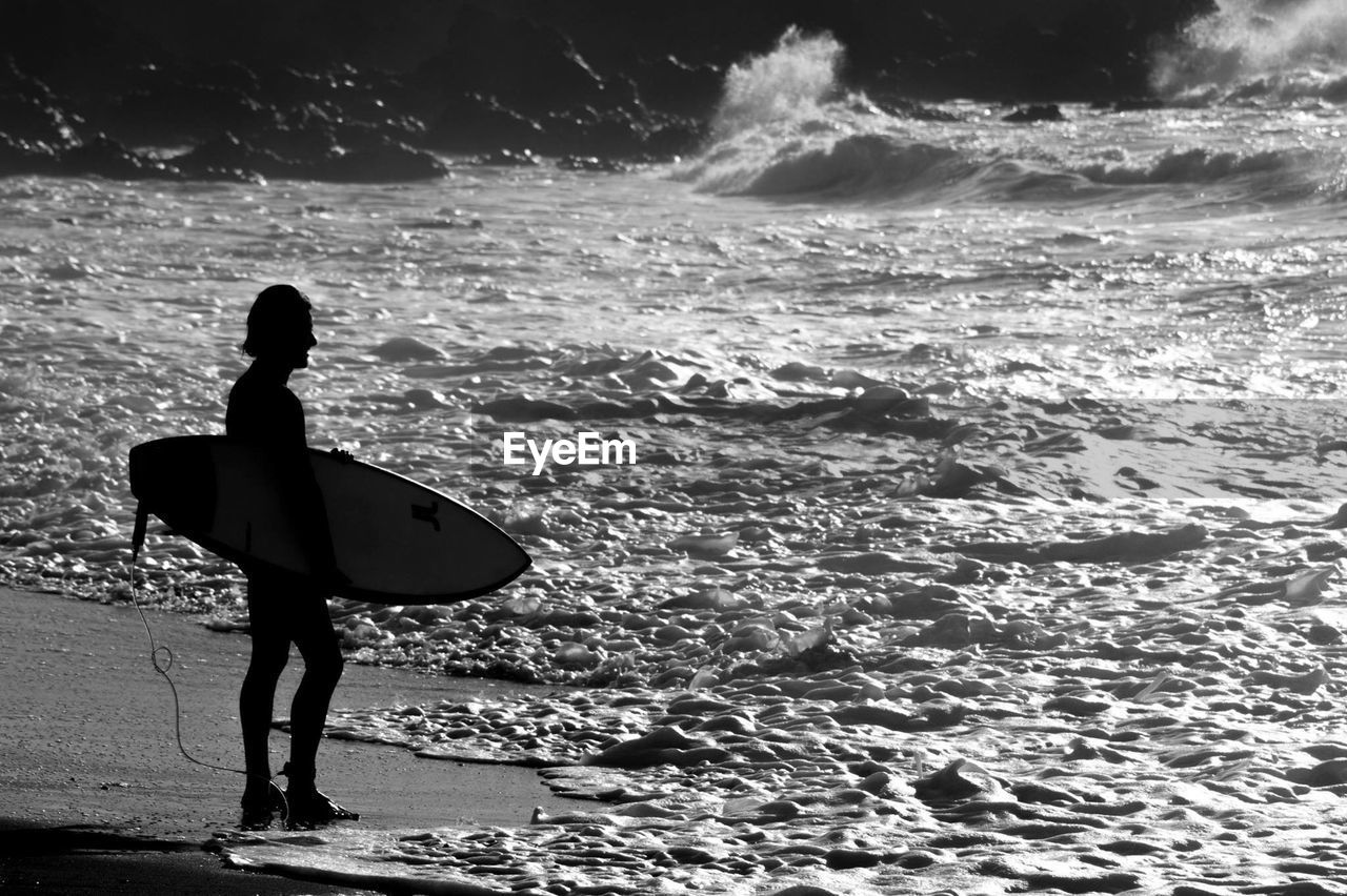 Full length of silhouette man with surfboard standing on shore at beach during sunny day