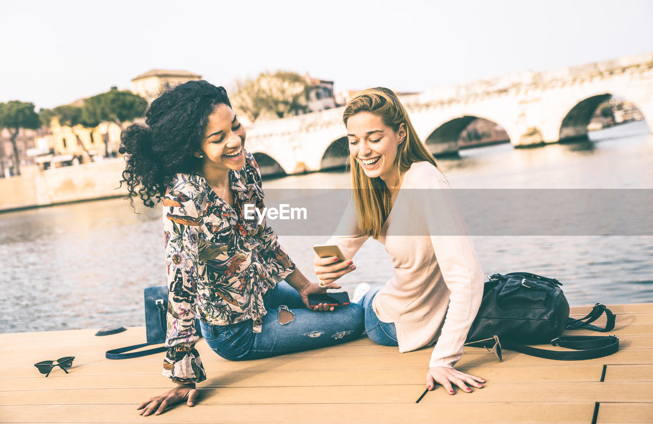 Female friends smiling while using mobile phone on pier over sea
