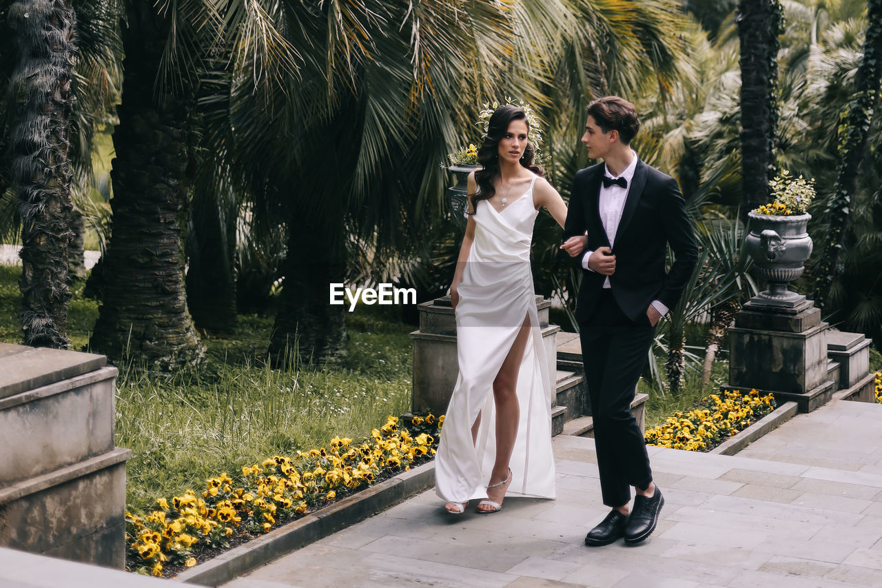 Happy lovers the bride and groom in wedding outfits walk among plants and palm trees in the old park