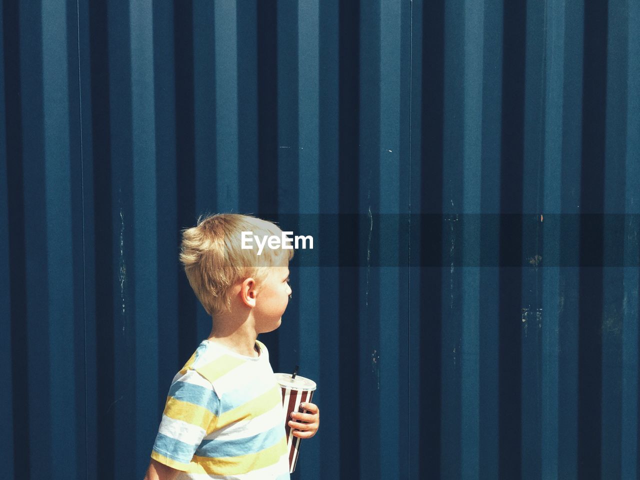 Side view of little boy standing against blue wall on sunny day