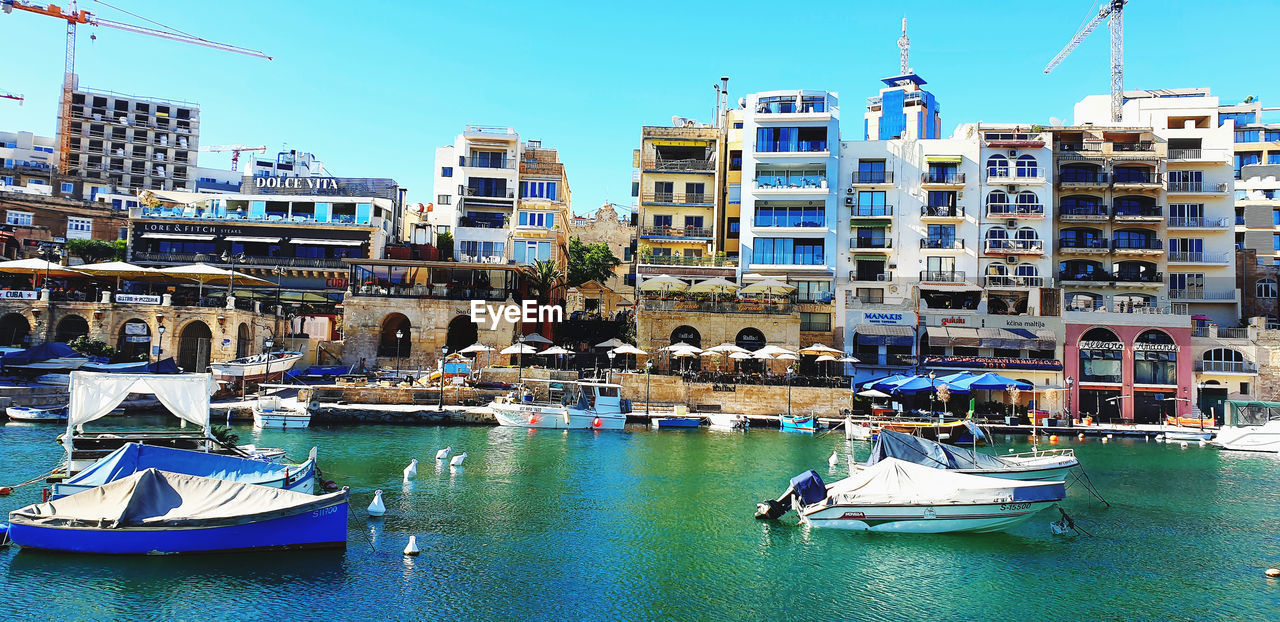 BOATS MOORED AT HARBOR BY BUILDINGS AGAINST SKY