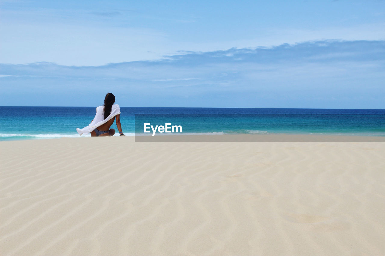 WOMAN SITTING ON BEACH AGAINST SKY