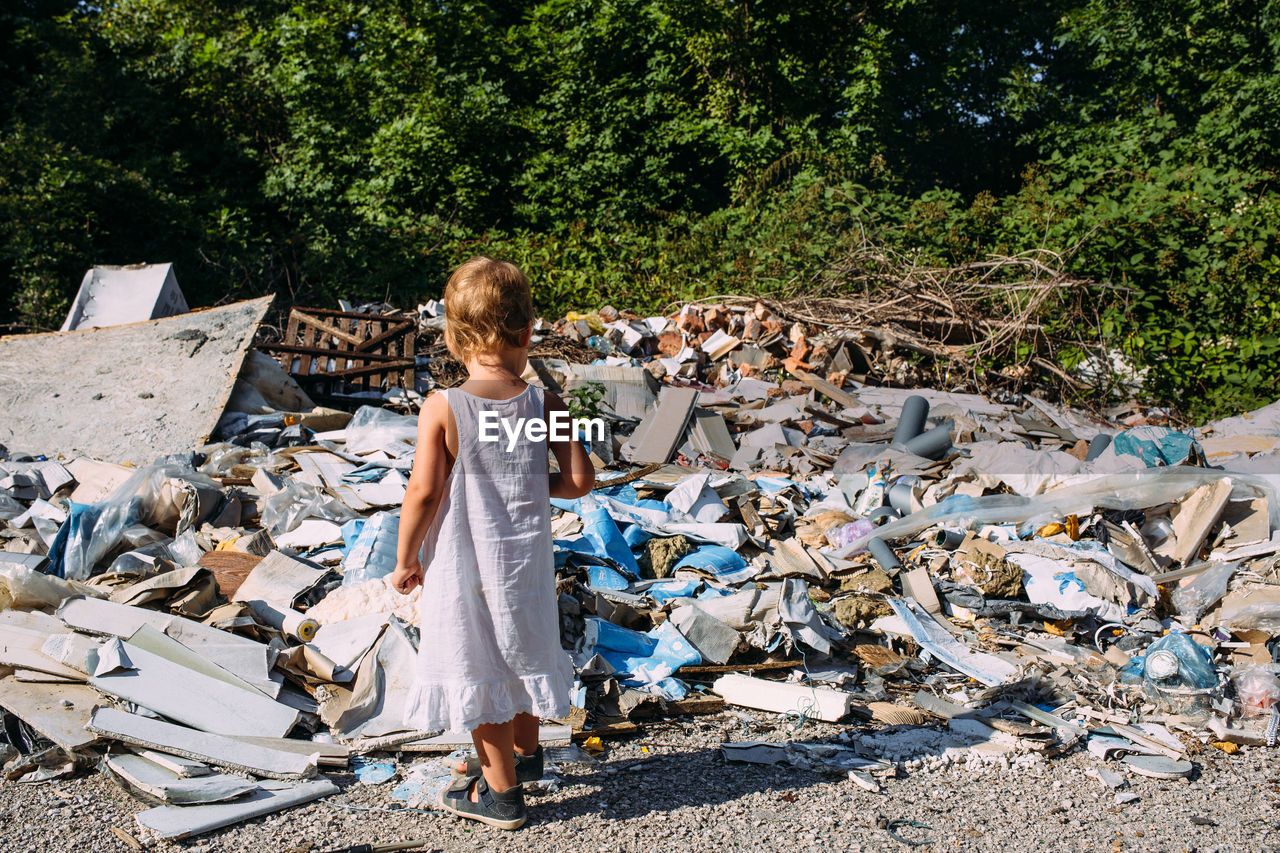 Little girl at a dump among a heap of scattered garbage in the forest.