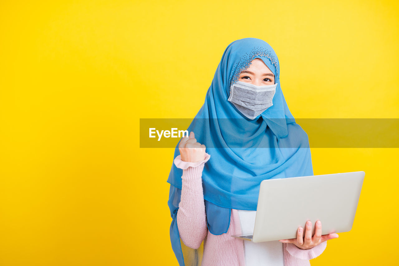 Young woman wearing mask holding laptop standing against yellow background