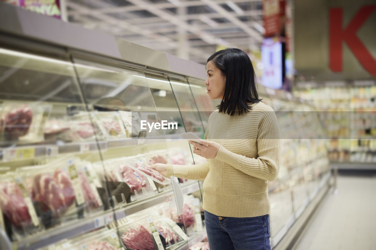 Woman doing shopping in supermarket and using cell phone to compare prices or checking shopping list