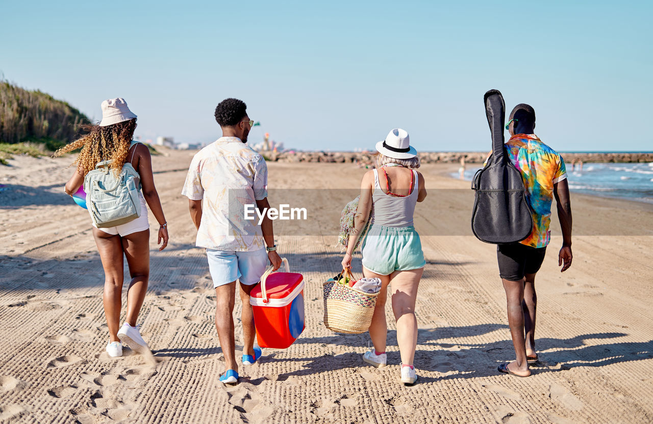 Full body back view of unrecognizable multiracial men and women in sunglasses and hat strolling on sand against cloudless blue sky while spending time on beach on summer day