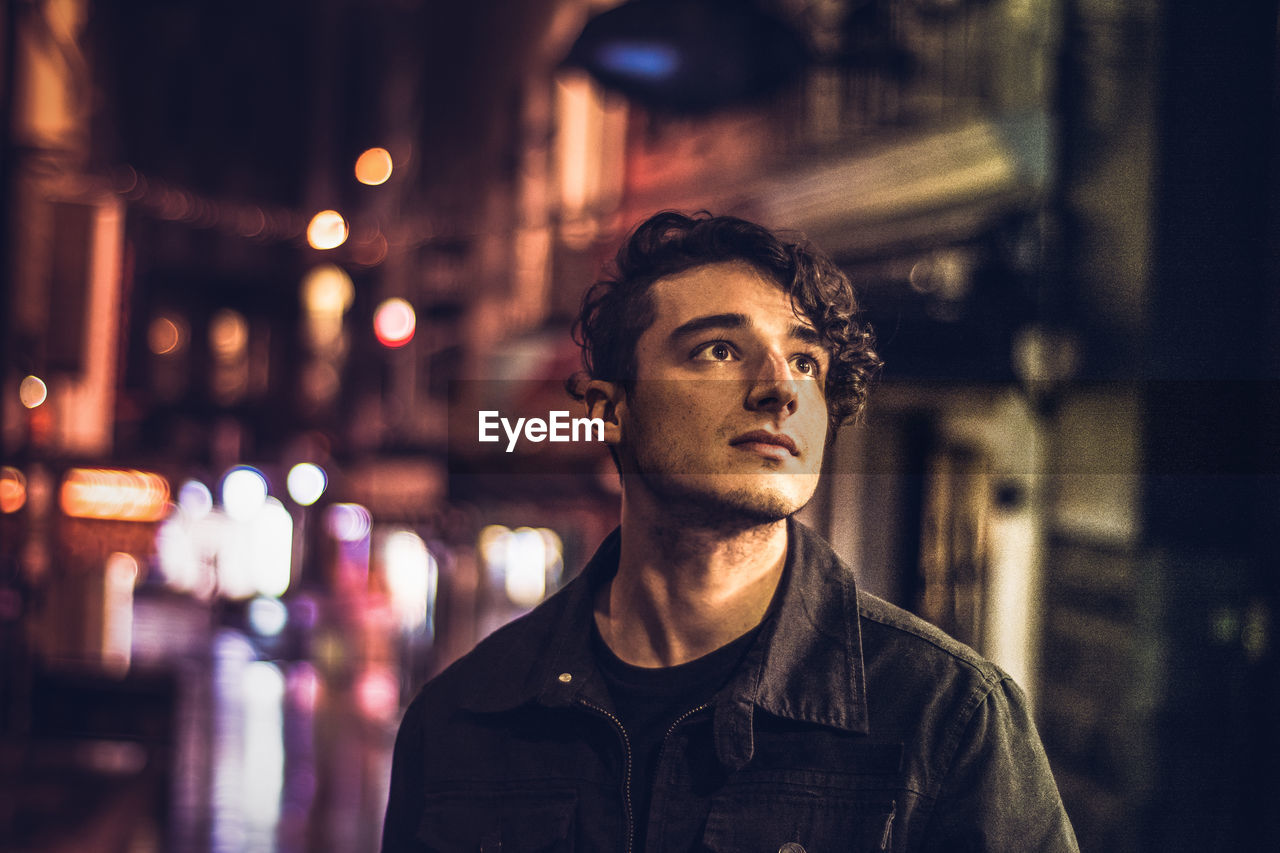 Thoughtful young man looking away while standing against buildings in city at night