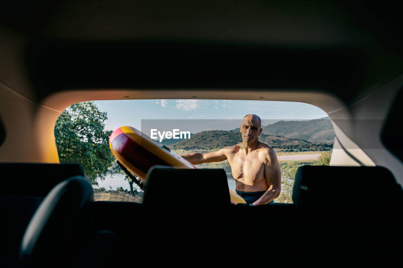 Active middle aged shirtless man taking paddle board from automobile parked on lake shore in hilly nature while preparing for water sport practice in summer day