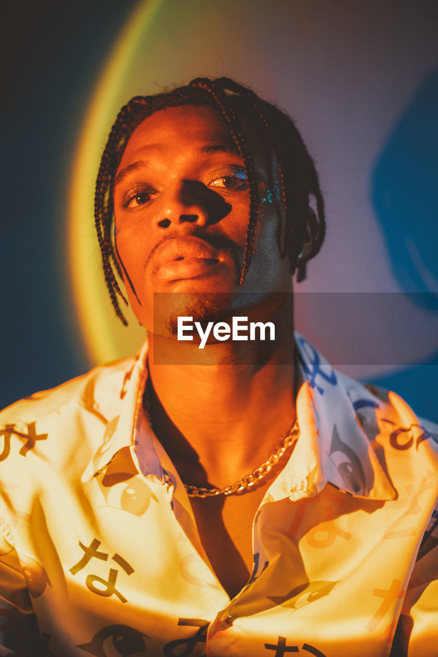 Serious african american male with braided hairstyle and chain looking at camera on dark background in studio with dim light