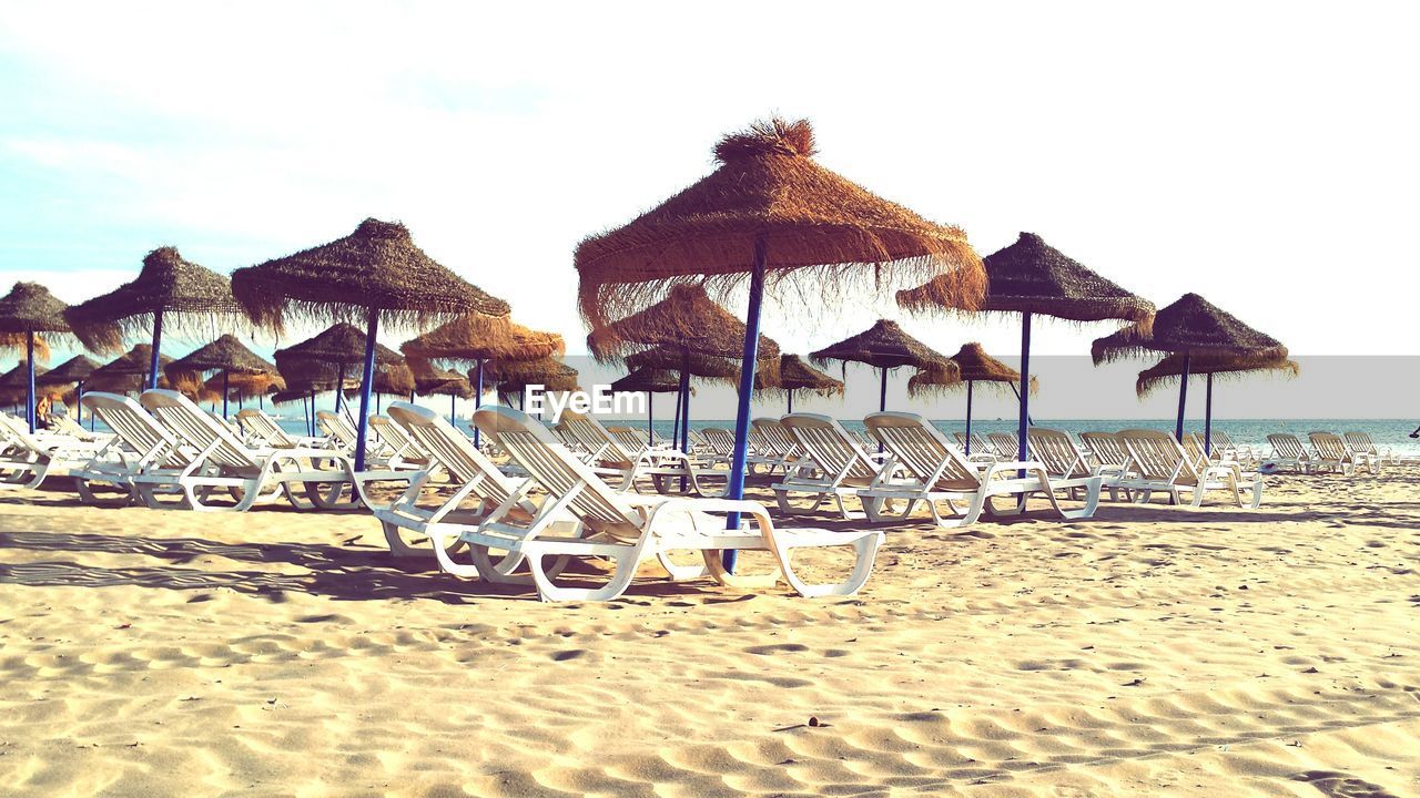 Thatched roofs and lounge chairs at beach against sky