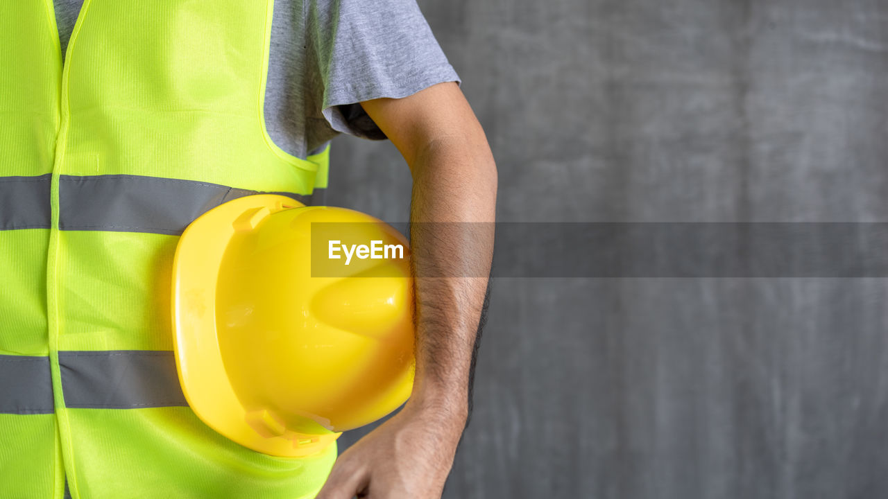 MIDSECTION OF MAN HOLDING YELLOW WHILE STANDING BY WALL