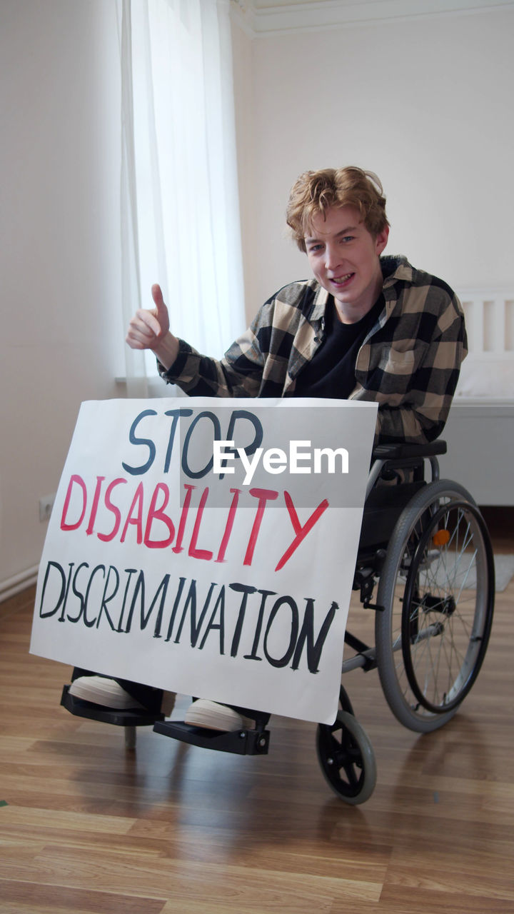 Portrait of smiling man holding poster sitting on wheelchair at home