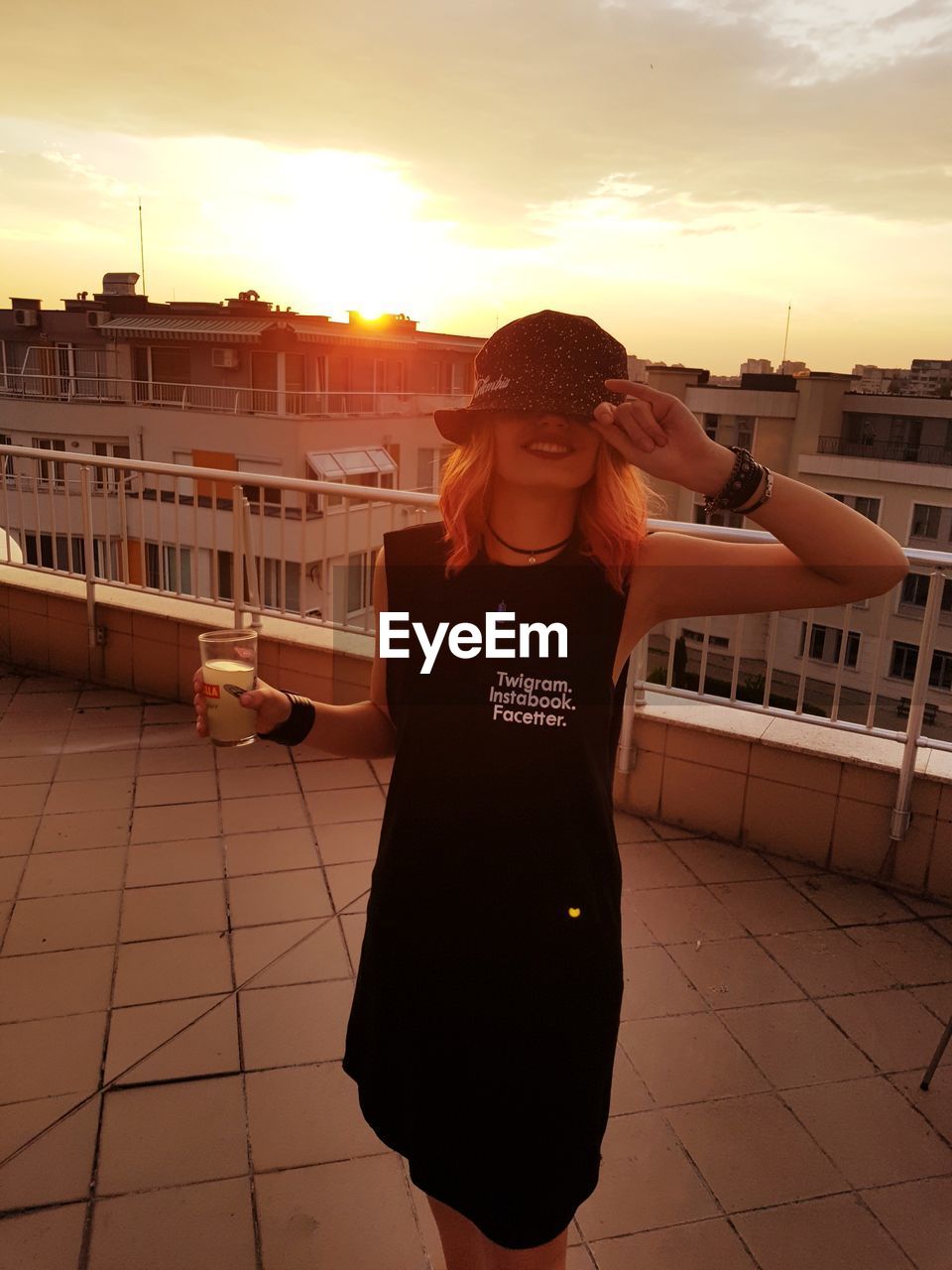 Woman holding drink while standing on terrace in city against sky