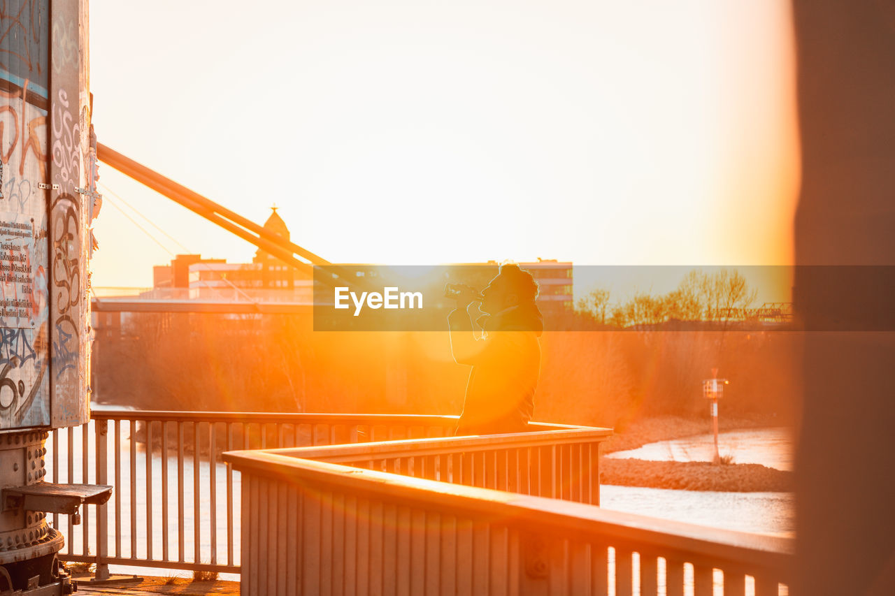 Rear view of man who is drinking beer and standing by railing against sky during sunset