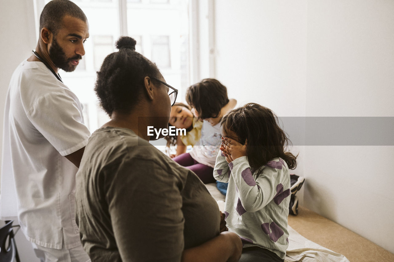 Male pediatrician and woman looking at girl covering eyes sitting with playful sisters