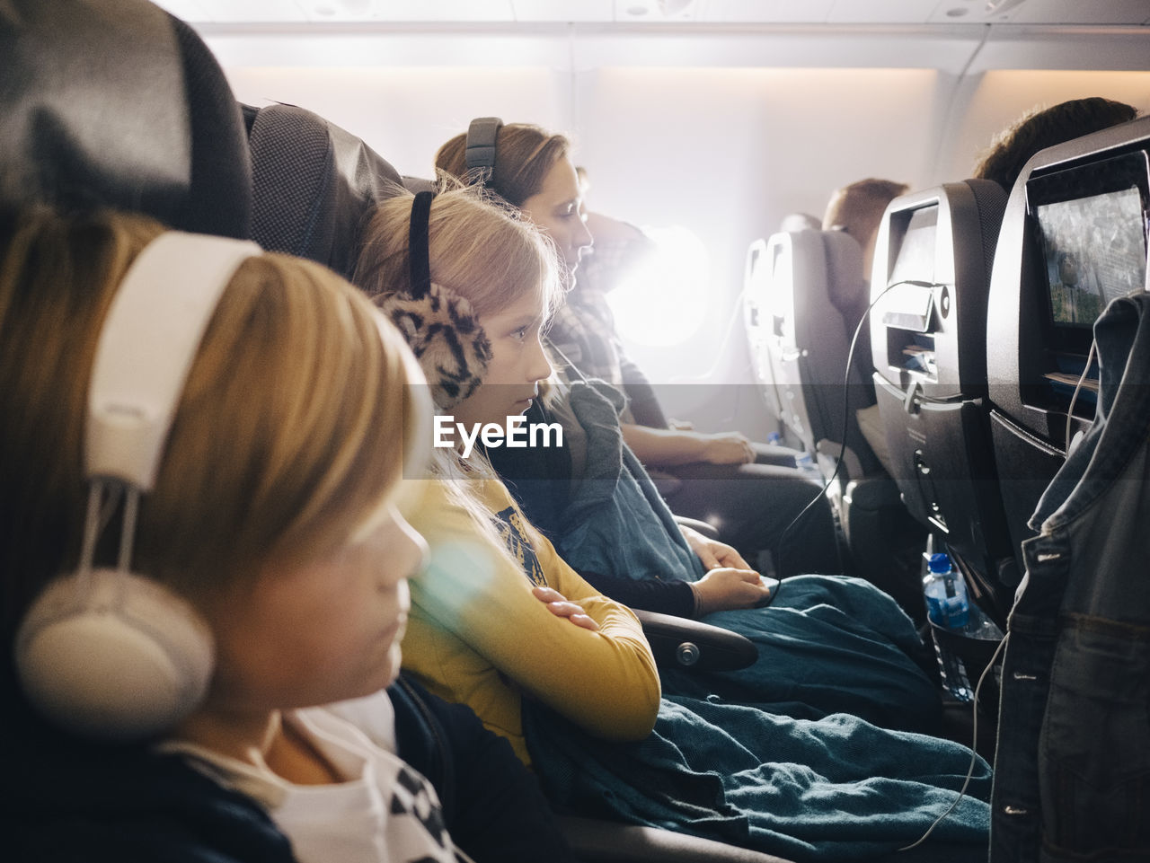 Side view of mother and children sitting in airplane while using headphones