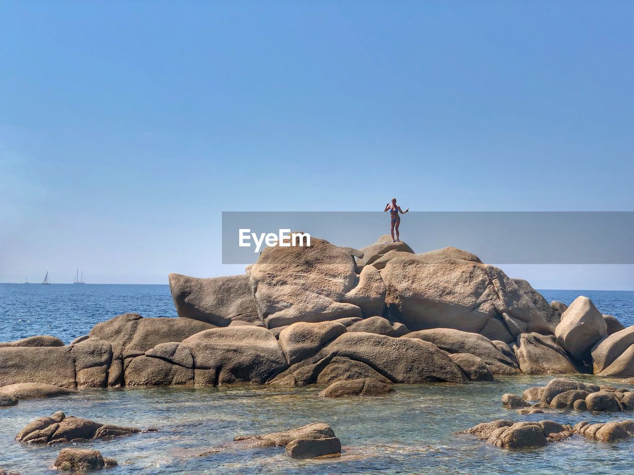 Distant view of woman standing on rocks in sea against clear blue sky