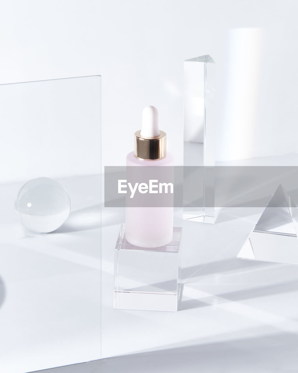 Glass bottle of pink hyaluronic acid for skin care treatment placed on table amidst glass geometric figures in light room