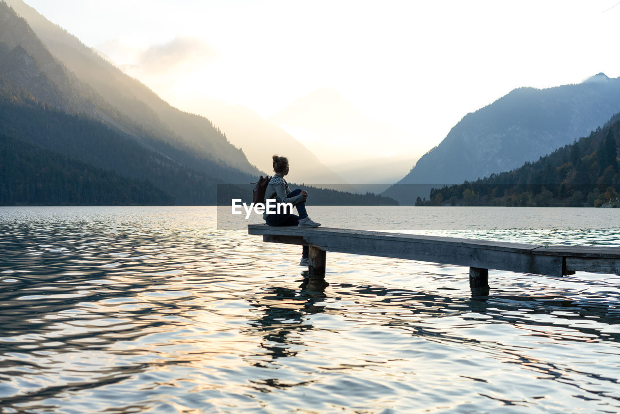 Side view of mid adult woman sitting on pier over lake by mountains