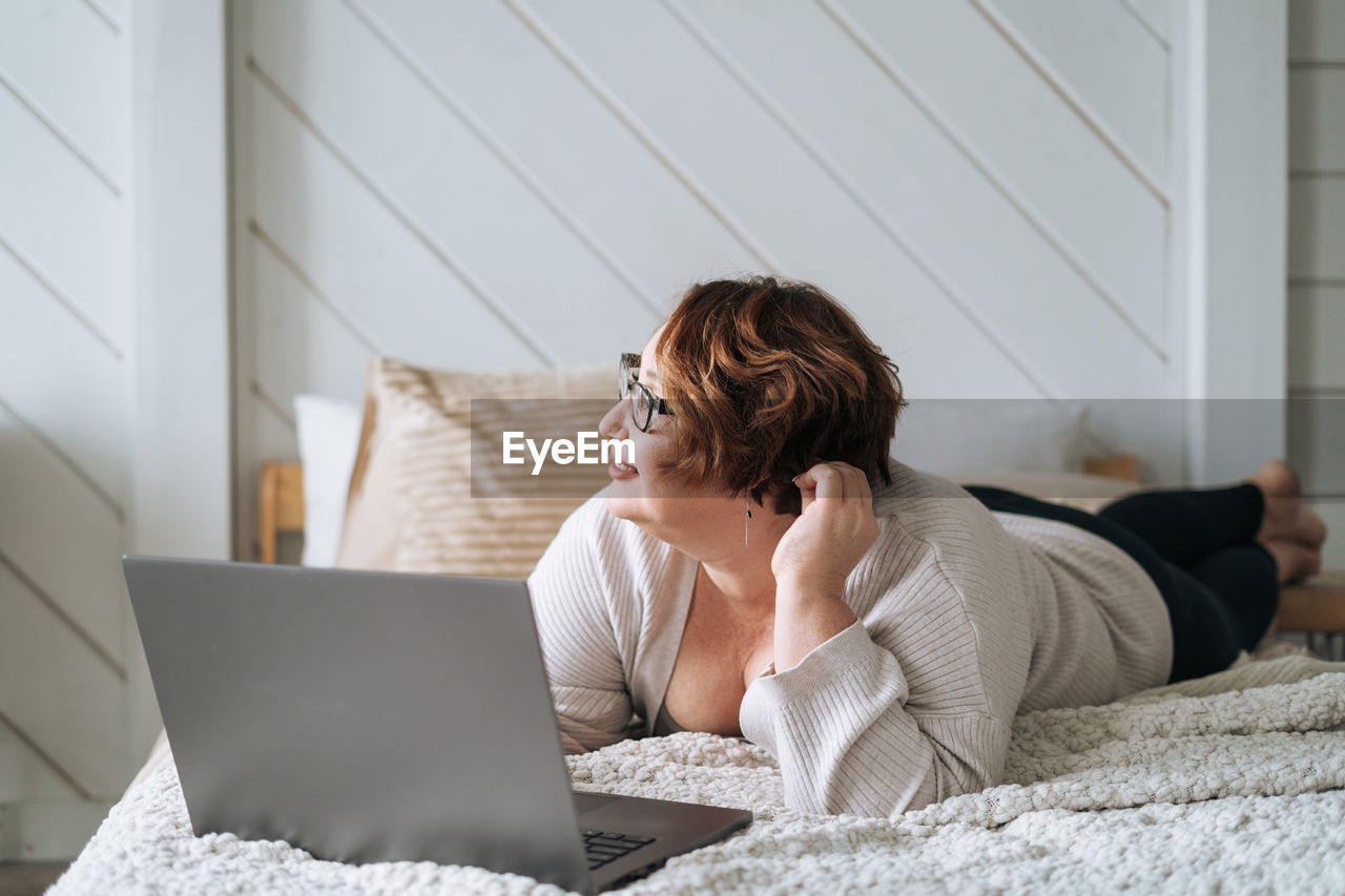 Smiling middle aged plus size woman lying on bed, working on laptop from home