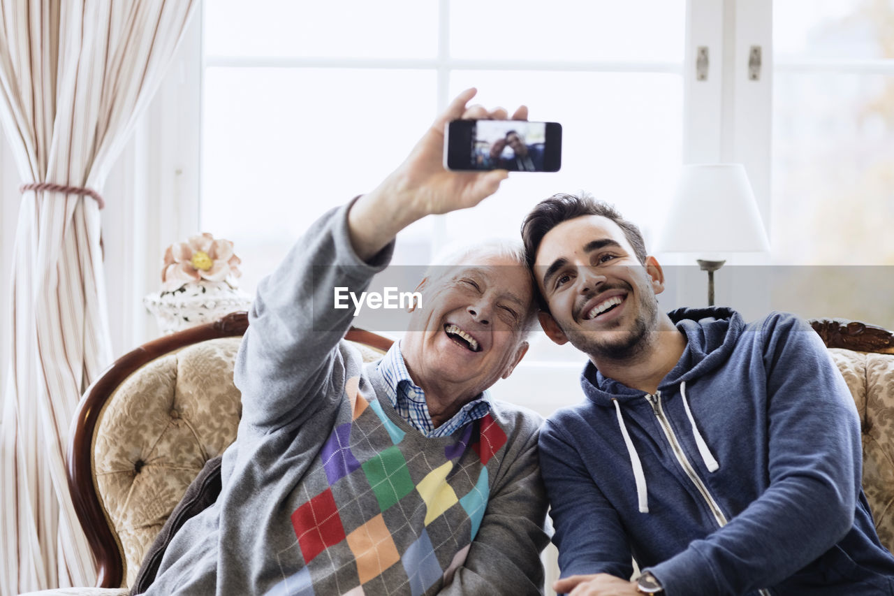 Happy senior man taking selfie with caretaker through smart phone at nursing home